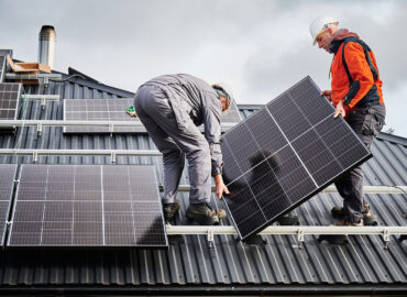 Engineers building solar panel system on roof of house. Men workers in helmets carrying photovoltaic solar module outdoors. Concept of alternative and renewable energy.