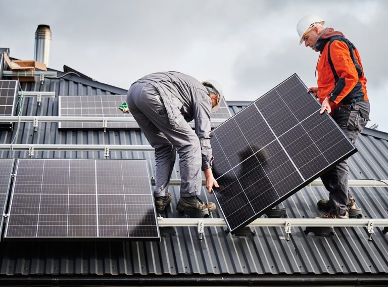 Engineers building solar panel system on roof of house. Men workers in helmets carrying photovoltaic solar module outdoors. Concept of alternative and renewable energy.